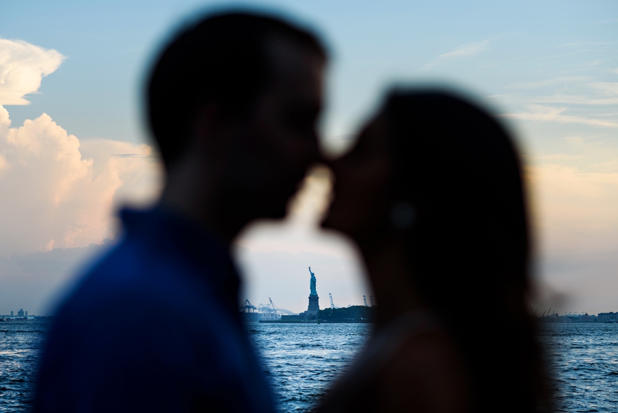 An engaged couple kiss with the statue of liberty in the background.